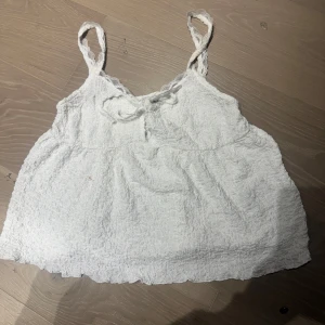 Vit spetsdetaljerad linnetopp - Söt vit linnetopp med spetsdetaljer på axelbanden och en liten rosett framtill. Toppen har en lätt skrynklig struktur och är ärmlös, perfekt för varma dagar.