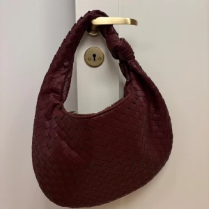 Design by si vinröd väska med knut - Säljer en vinröd axelväska med en snygg knut på handtaget. Väskan har dragkedja upptill och är rymlig med ett mjukt, rundat formspråk. Perfekt accessoar för att lyfta din outfit. Använd en gång. 