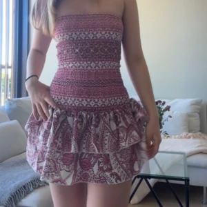 Rosa och vit axelbandslös kortklänning med volang - Supersöt axelbandslös kortklänning i rosa och vitt med smockad överdel och volangkjol. Klänningen har ett sommrigt mönster och sitter tight upptill med en utställd kjol. Perfekt för varma dagar! Inbyggda shorts! 