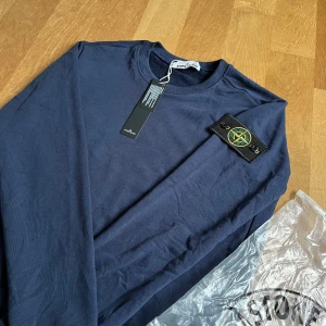 Mörkblå långärmad tröja från Stone Island - Säljer en mörkblå långärmad tröja från Stone Island med den klassiska logotypen på ärmen. Tröjan har rund halsringning och är tillverkad i mjukt material. Perfekt för en stilren och avslappnad look.
