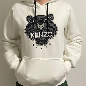 Vit hoodie från Kenzo med tigertryck - Säljer en vit hoodie från Kenzo med stort svart tigertryck och logga på bröstet. Tröjan har huva med svarta snören och en stor magficka. Tillverkad i mjuk bomull. OBS! Tröjan är i storlek XL men har passform för M.