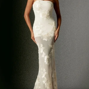 Balklänning  - Strl M men passar S. Elegant axelbandslös vit långklänning med transparenta partier och stora blommiga spetsapplikationer. Klänningen har en figurnära passform och en lätt släp baktill, perfekt för speciella tillfällen.