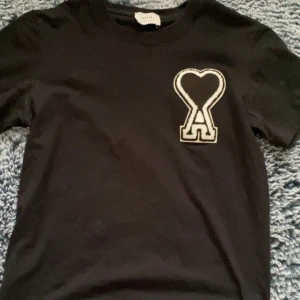 Svart t-shirt med hjärtdetalj från Ami - Svart t-shirt från Ami med en stor vit broderad hjärtdetalj och bokstaven A på bröstet. Klassisk rund hals och korta ärmar. Perfekt för en stilren och trendig look.