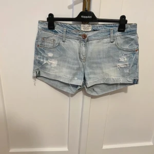 Ljusblå lågmidjade jeansshorts från River Island - Midjemått 44 cm. Säljer ett par ljusblå jeansshorts från River Island med slitna detaljer och fransar. Shortsen har klassisk femficksmodell, knappgylf och bälteshällor. Perfekta för varma dagar och en avslappnad stil. Superfina och lågmidjade!