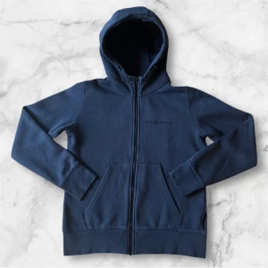 Svart hoodie från Sail Racing - Säljer en mörkblå hoodie från Sail Racing med dragkedja och två stora fickor framtill. Tröjan har huva och diskret logga på bröstet. Perfekt för en avslappnad stil och enkel att matcha med det mesta.