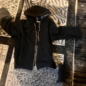 Svart hoodiejacka från Burberry - Svart hoodiejacka från Burberry med dragkedja och klassiskt rutigt foder i huvan. Jackan har två fickor framtill och en stor huva. Perfekt för en stilren och avslappnad look.