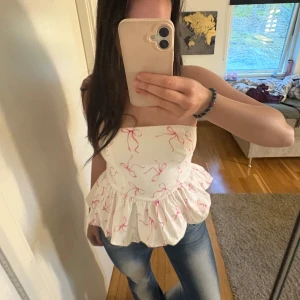 Topp  - Supersöt axelbandslös topp i vitt med rosa rosetter och volang nertill. Perfekt för sommardagar och ger en romantisk vibe till outfiten. Säljs då den inte kommer till användning längre.