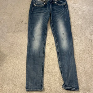 Blå jeansbyxor med nitar från Miss Me - Snygga blå jeansbyxor från Miss Me med dekorativa nitar och stenar på fickorna. Byxorna har en klassisk femficksmodell och normal passform. Perfekta för dig som vill sticka ut med detaljerade jeans! Storlek 30 och det står skinny men det är mycket mer straight leg på dessa 
