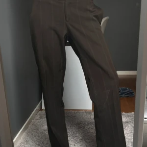 Bruna kostymbyxor med diskreta ränder - Snygga bruna kostymbyxor med subtila vertikala ränder och klassisk passform. Byxorna har dragkedja och knapp framtill och är perfekta för en stilren look. Nertill så har jag ”sprättat upp” sömmen, för att förlänga benen. Förra ägaren har sytt om runt skrevet för att byxorna ska vara lågmidjade! Mycket snyggt 