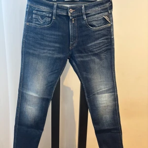 Replay Anbass jeans i blå denim - Säljer ett par Replay Anbass jeans i klassisk blå tvätt med snygga slitningar. Jeansen har fem fickor, dragkedjegylf och smal passform. Tillverkade i hållbar bomull för en bekväm känsla. Perfekta för dig som gillar stilrena och moderna jeans.