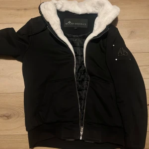 Svart Moose Knuckles jacka - Säljer en svart jacka från Moose Knuckles med vit fluffig huva och quiltat innerfoder. Jackan har dragkedja framtill och en logotyp i metall på ärmen. Perfekt för kyligare dagar och har en modern look.
