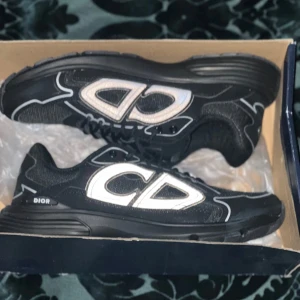 Dior b30 - Säljer ett par svarta sneakers från Dior med stora vita CD-loggor på sidorna. Skorna har en sportig design med platt sula och snörning. Perfekta för dig som vill sticka ut med en lyxig touch. Aldrig använd fel storlek tveka inte på att skriva till mig