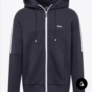 Marinblå hoodiejacka med dragkedja från BOSS - Snygg marinblå hoodiejacka från BOSS med vit dragkedja och smala vita ränder längs ärmarna. Jackan har huva med snörning och två fickor framtill. Perfekt för en avslappnad och sportig stil.
