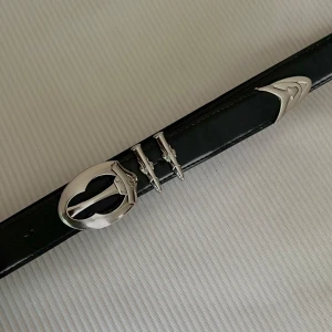 Avant-Garde Noir Belt - Svart bälte med unika silverdetaljer som ger en avantgardistisk och stilren känsla. Perfekt för dig som vill addera en modern och uttrycksfull touch till din look. Första hålet vid 66,5 cm, sista vid 89 cm. Total längd: 101 cm
