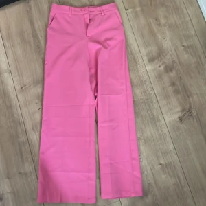 Rosa kostymbyxor från Lindex - Säljer ett par snygga rosa kostymbyxor från Lindex med raka, vida ben och hög midja. Byxorna har hällor för bälte, knapp och dragkedja framtill. Perfekta för dig som vill sticka ut med färg!