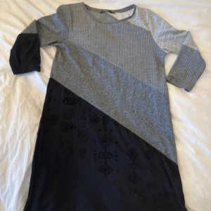 Grå och svart kortklänning Desigual - Snygg kortklänning från Desigual med diagonala färgblock i grått och svart. Klänningen har trekvartsärm, rund hals och coolt geometriskt mönster på den svarta delen. Materialet är mjukt och stretchigt, perfekt för dig som gillar bekväma och stilrena plagg.