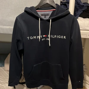 Mörkblå hoodie Tommy Hilfiger M - Snygg mörkblå hoodie från Tommy Hilfiger med vit logotyp och broderad flagga på bröstet. Tröjan har huva med vita snören, stor magficka och långa ärmar. Perfekt för chill dagar. Klassisk design med coola detaljer.