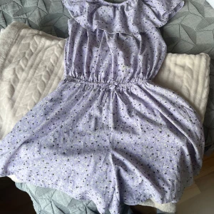 Ljuslila blommig kortklänning Basic Needs - Supersöt ljuslila kortklänning från Basic Needs med små blommor i lila och gult. Klänningen har volang upptill och resår i midjan för en skön passform. Perfekt för sommarens alla dagar, lätt och luftig i materialet.