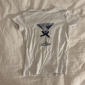 Vit baby tee med martiniglas från Subdued - Vit t-shirt från Subdued i bomull med ett stort tryck av ett martiniglas och en mörkblå rosett på framsidan. Modellen är kortärmad och har en klassisk rund hals. Perfekt för dig som gillar coola och unika prints.