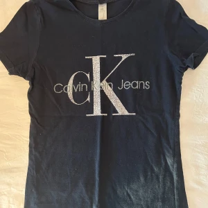 Svart Calvin Klein Jeans t-shirt - Svart t-shirt från Calvin Klein Jeans med stort CK-tryck i silver på bröstet. Klassisk rund hals och korta ärmar. Materialet är mjuk bomull som sitter skönt mot kroppen. Perfekt för dig som gillar stilrena logotoplagg och vill ha en enkel men snygg look. Ganska bra som sovtröja