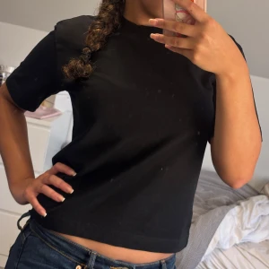 Svart basic tröja från Gina Tricot - En svart, enkel och stilren t-shirt från Gina Tricot, den är lite tjockare än vanliga T-shirts. Den e från basically basics-kollektionen. Tröjan har rund hals, korta ärmar och är gjord i mjuk bomull. Perfekt till jeans eller under en jacka. Snygg passform som sitter normalt och passar till många olika looks.