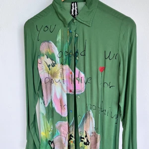 Grön skjorta med blommor från Desigual - Unik grön skjorta från Desigual med handmålat blommönster i rosa och beige på framsidan. Skjortan har tryckt text, små broderade detaljer och knappar framtill. Långärmad och i mjukt, tunt material som känns riktigt skönt mot huden.