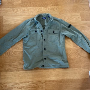 Grön overshirt från Jack & Jones - Snygg militärgrön overshirt från Jack & Jones med svarta knappar och två bröstfickor med lock. Skjortan är i bomull och har lång ärm, perfekt att slänga över en t-shirt för en chill look.