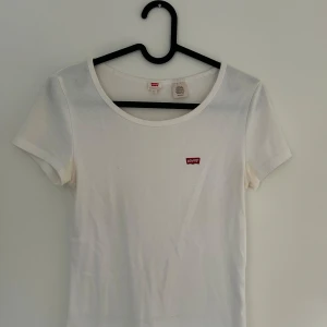 Vit t-shirt från Levi's med logga - Vit t-shirt från Levi's med klassisk rund hals och korta ärmar. På bröstet sitter den ikoniska röda Levi's-loggan som ger en clean och stilren vibe. Perfekt till jeans eller shorts för en enkel och vardaglig look. Nästintill oanvänd.