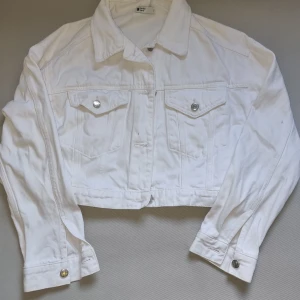 Vit croppad jeansjacka från Gina Tricot - Snygg vit croppad jeansjacka från Gina Tricot med klassisk krage, silverfärgade knappar och två bröstfickor med lock. Jackan har långa ärmar och är tillverkad i bomull. Perfekt för att lyfta vilken outfit som helst.