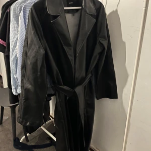 Svart lång skinn trenchcoat Boohoo - Säljer en svart lång trenchcoat i skinnimitation från Boohoo. Jackan har klassisk krage, bälte i midjan och raka ärmar. Passformen är loose och passar också 38 