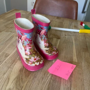 Blommiga gummistövlar med kaninmotiv - Söta gummistövlar från Next i storlek 22 med färgglatt blommönster och gulligt kanintryck. Stövlarna har rosa detaljer och praktiska dragflikar upptill. Perfekta för regniga dagar och lek utomhus.