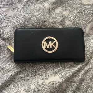 Svart plånbok från Michael Kors - Snygg svart plånbok från Michael Kors med stort guldfärgat MK-emblem framtill. Plånboken har dragkedja runtom, flera kortfack och ett innerfack med dragkedja. Tillverkad i strukturerat skinn med gulddetaljer och klassisk, stilren design.