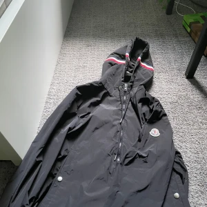 Äkta Svart Moncler vindjacka med huva - Svart vindjacka från Moncler med huva och dragkedja framtill. Jackan har Moncler-logga på armen och snygga ränder i vitt och rött på huvan. Tillverkad i lätt polyester, perfekt för blåsiga dagar. Fickor med tryckknappar och stilren design. Den har även nfc scan den passar mig bra som är 190cm men passar dig även som är runt 180