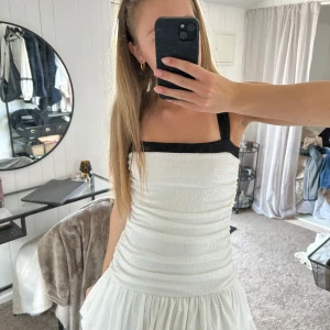 Vit kort klänning med svarta axelband - Supersöt vit kort klänning med svarta breda axelband och fyrkantig urringning. Klänningen har rynkad överdel och voluminös kjol med volang. Perfekt för dig som gillar stilrena kontraster och trendiga detaljer. Passar även S. Använd 1 gång. 