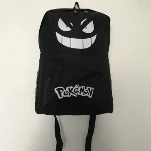 Svart Pokémon ryggsäck  - Cool svart ryggsäck med stort vitt Gengar-ansikte och Pokémon-logga framtill. Rymligt huvudfack, ytterficka med dragkedja och meshfickor på sidorna. Justerbara axelremmar och slitstarkt material – perfekt för skolan eller häng med kompisar.