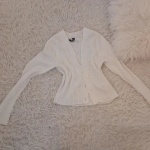 Vit ribbad kofta från H&M Divided - Säljer en vit ribbad kofta från H&M Divided i storlek S. Koftan har lång ärm, v-ringad hals och tre stora knappar framtill. Den är figurnära och gjord i mjukt stickat material, perfekt för lager-på-lager under hösten och vintern.