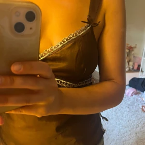 Brun omlott linnetopp med spets - Snygg brun omlott linnetopp med tunna axelband och dekorativ spetskant i vitt längs halsringningen och under bysten. Toppen har rosett-detaljer på axelbanden och sidorna, vilket ger en extra söt touch. Materialet är glansigt och känns lyxigt mot huden.