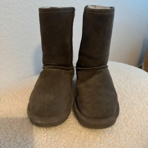 Gråa Bearpaw boots i mocka - Säljer ett par gråa Bearpaw boots med rund tå och platt sula. Skorna är tillverkade i mjuk mocka och har en klassisk, enkel design med högt skaft. Insidan är fodrad för extra värme, perfekt för kyliga dagar. Märkeslogga bak på hälen. Använd en gång. Köpta i USA.