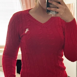 Röd stickad tröja från Ralph Lauren - Säljer en snygg röd stickad tröja från Ralph Lauren med klassisk kabelstickning och vit broderad logga på bröstet. Tröjan har v-ringad hals och långa ärmar, perfekt för dig som gillar preppy stil och vill ha något som sticker ut.