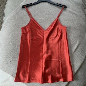 Röd satinlinne med smala axelband XS - Säljer ett rött linne i satin med smala axelband och v-ringning. Toppen har en slät, glansig yta och en enkel, stilren design som känns både trendig och klassisk. Perfekt att styla med jeans eller kjol för en snygg look.