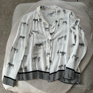 Vit zebramönstrad skjorta från MNG - Snygg vit skjorta från MNG med svart zebramönster över hela plagget. Skjortan har långa ärmar, klassisk krage och svarta ränder vid ärmslut och nederkant. Tillverkad i mjuk och lätt polyester, perfekt för dig som gillar djurprint och vill sticka ut.