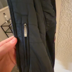 Svart fleecejacka Parajumpers S - Svart fleecejacka från Parajumpers i storlek S. Jackan har quiltade detaljer, dragkedjor och en snygg patch på ärmen. Insidan är ljusgrå och materialet är mjukt fleece. Perfekt för dig som vill ha en stilren och funktionell jacka med coola detaljer.