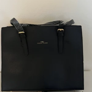 Svart handväska från Tnig Concept Milano - Stilren svart handväska med rektangulär form och guldfärgade detaljer. Väskan har justerbara handtag och medföljande axelrem. Tillverkad i skinnimitation med slät yta och dragkedja upptill. Perfekt för att bära med sig allt viktigt.OBS väskan är från zalando och aldrig använd.