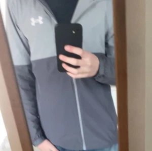 Grå vindjacka från Under Armour - Snygg tvåfärgad grå vindjacka från Under Armour med dragkedja framtill och elastiska muddar vid ärmsluten. Jackan har en sportig look och är perfekt för blåsiga dagar. Loggan syns tydligt på bröstet och materialet känns lätt och smidigt. 9/10 skick!