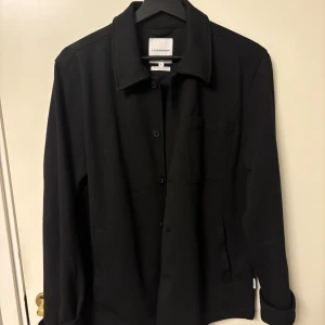 Svart overshirt från Lindbergh - Stilren svart overshirt från Lindbergh med klassisk krage, knäppning framtill och bröstficka. Perfekt lager-på-lager-plagg med enkel design och raka linjer. Tillverkad i mjukt och stretchigt material. Använd ungefär 5 gånger finns inga tecken på användning. Passar s-m