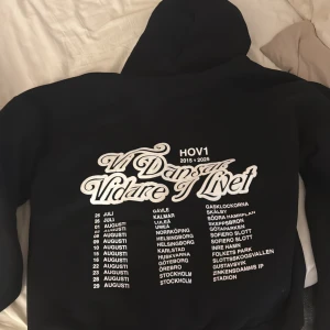 Svart HOV1 hoodie med turnétryck - Svart hoodie från HOV1 med stor vit text på ryggen: 'Vi Dansar Vidare I Livet' och turnédatum samt städer. Klassisk huva och långärmad modell, perfekt för dig som är hov1 fan och vill ha merch till minne av dem! 
