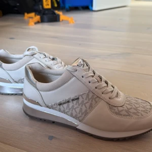 Beige och silver sneakers Michael Kors - Snygga sneakers från Michael Kors i beige och silver med logomönster på sidan. Skorna har detaljer i skinn och textil, samt metallic-silver på hälen och sula. Klassisk snörning och platt sula gör dem både stilrena och bekväma. Perfekta för dig som gillar lyxig streetstyle.