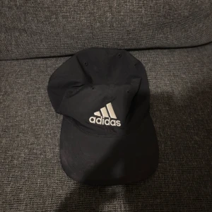 Svart keps från Adidas - Svart keps från Adidas med klassisk böjd skärm och broderad vit logga framtill. Justerbar baktill för perfekt passform. Tillverkad i bomull, perfekt för en sportig och avslappnad look.