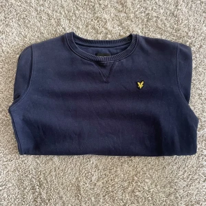 Mörkblå sweatshirt från Lyle & Scott - Snygg mörkblå sweatshirt från Lyle & Scott Junior med klassisk rund hals och ribbade muddar. Tröjan har ett gult broderat logomärke på bröstet och är tillverkad i mjuk bomull. Perfekt för dig som gillar stilrena och bekväma plagg.