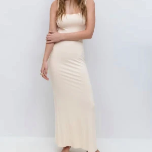 Champagnefärgad långklänning med smala axelband slip dress Gina - Säljer en stilren och elegant långklänning i champagnefärg med smala axelband och figurnära passform. Klänningen har en enkel, rak siluett och är ärmlös, vilket ger en modern och minimalistisk look. Perfekt för dig som gillar en clean och chic stil.